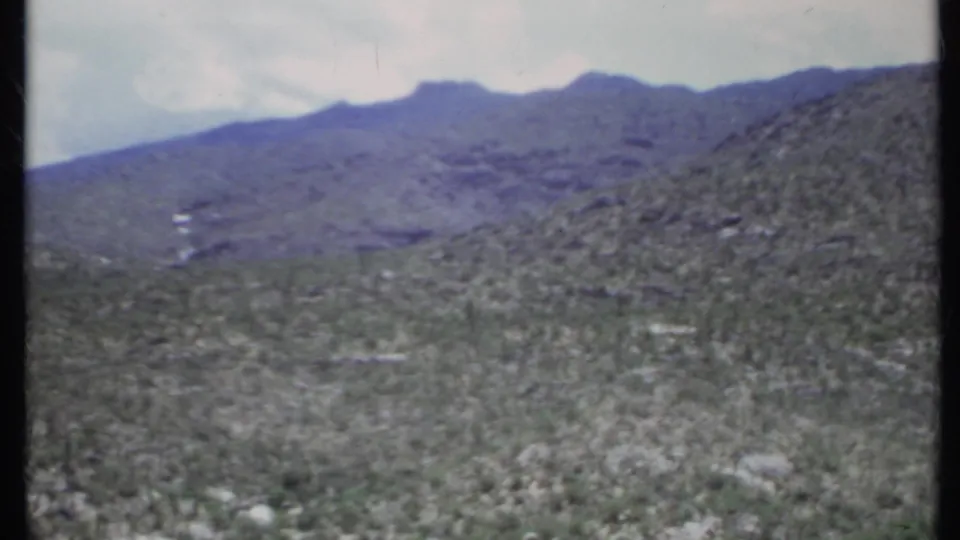 1971 California desert view with scrub-covered ground, rocky hillside, and blue-gray mountains softened by 8mm grain and blur.