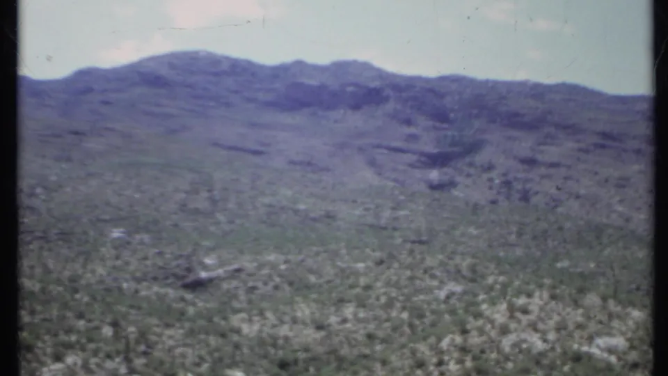 Home-movie frame from 1971 California showing an open rocky desert slope, scattered low vegetation, and distant mountains under hazy light.