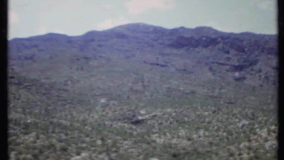 1971 California 8mm frame of a broad arid hillside, scattered scrub, and a distant rocky ridge with soft focus and muted color.