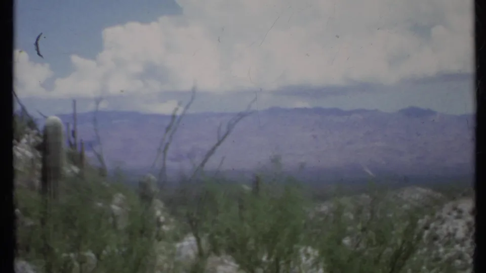 1971 8mm California desert scene with cactus at left, scrub foreground, hazy mountains, and sunlit white clouds.