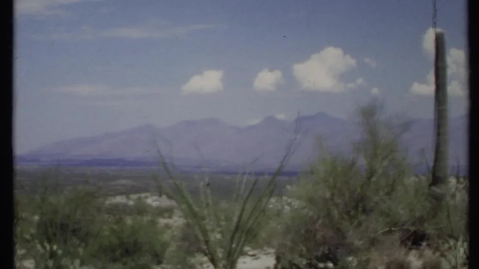 1971 8mm frame showing cactus, scrub, dry desert terrain, purple-hazed mountains, and small white clouds.