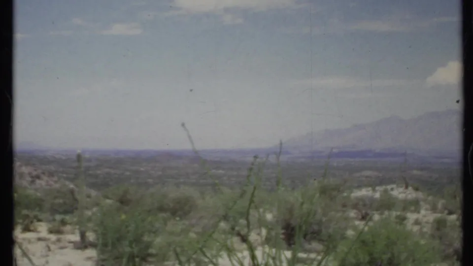 Wide 1971 home-movie desert vista with sparse shrubs, hazy mountain forms, pale ground, and washed blue sky.