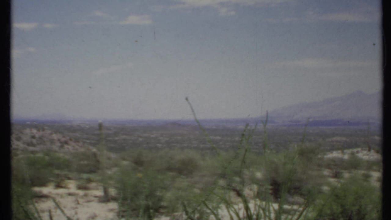 1971 California 8mm view of sparse desert vegetation and sandy ground, with hazy mountains on the horizon.