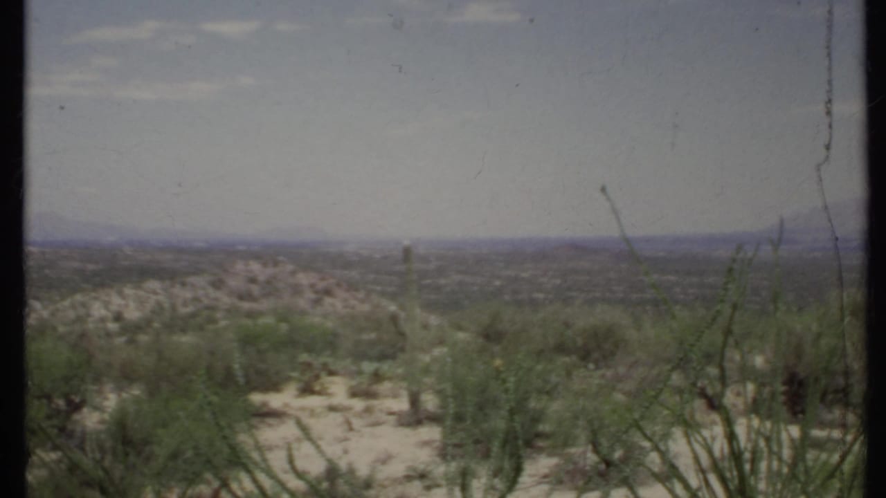 Open desert plain with scrub vegetation, low hills, and a hazy mountain horizon.