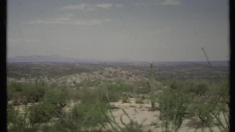 Vintage 8mm footage frames a sandy desert opening among shrubs, looking toward low hills and a distant mountain line.