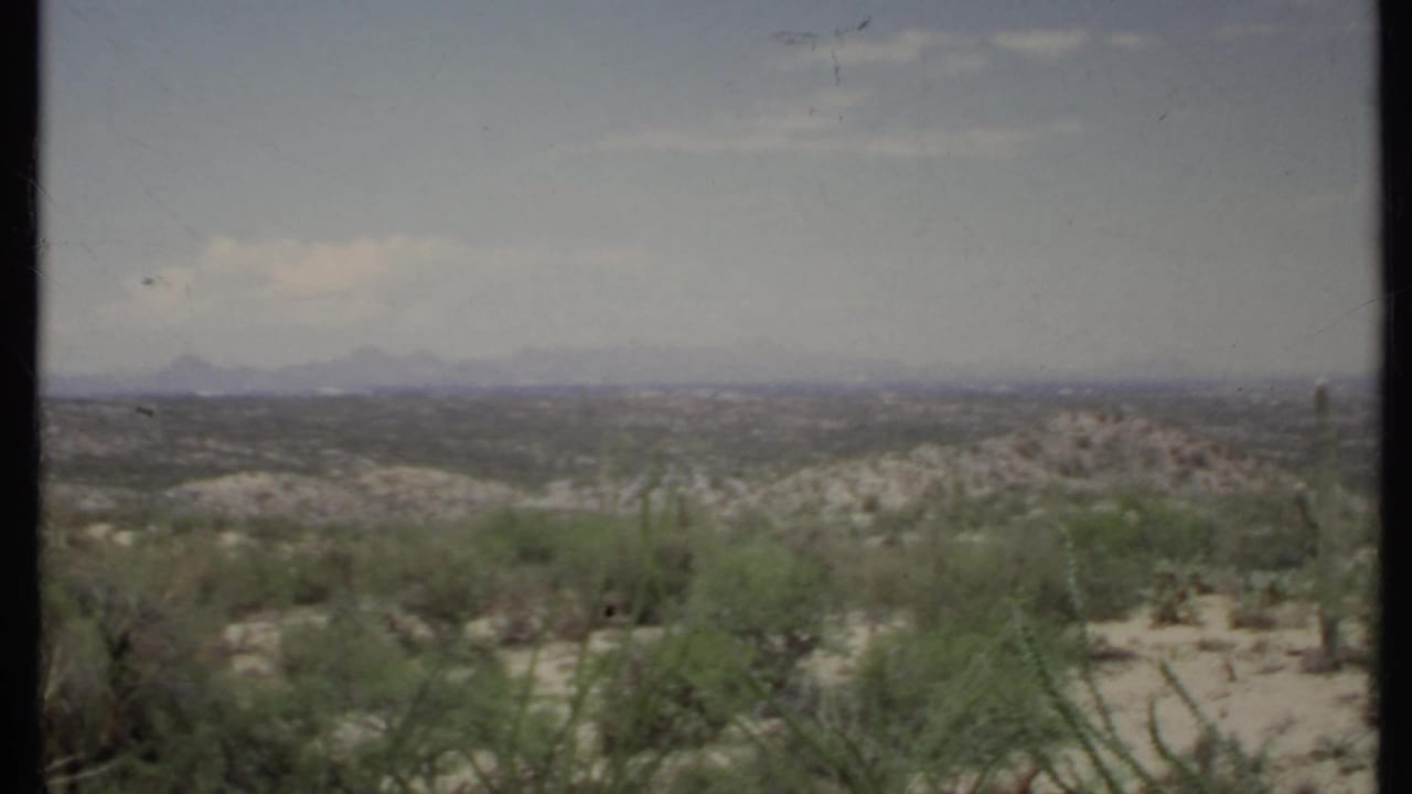 1971 California 8mm frame showing open desert shrubs, low sandy ground, distant ridges, and a lightly clouded blue sky.