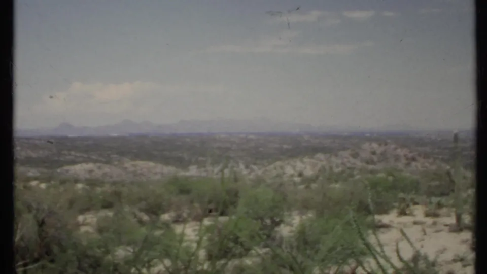 1971 California 8mm frame showing open desert shrubs, low sandy ground, distant ridges, and a lightly clouded blue sky.
