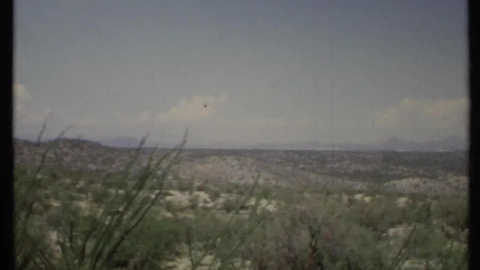 1971 California 8mm frame showing near desert plants, sandy washes, low hills, and faint mountains under a pale sky.