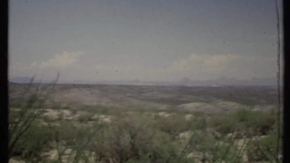1971 California 8mm frame of open desert shrubland, rolling hills, hazy mountain forms, and broad sky with soft clouds.