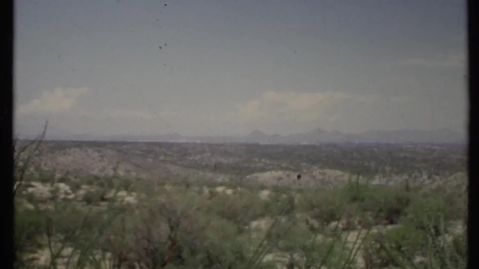 1971 8mm California desert view with brushy foreground, open valley, and faint mountain silhouettes on the horizon.