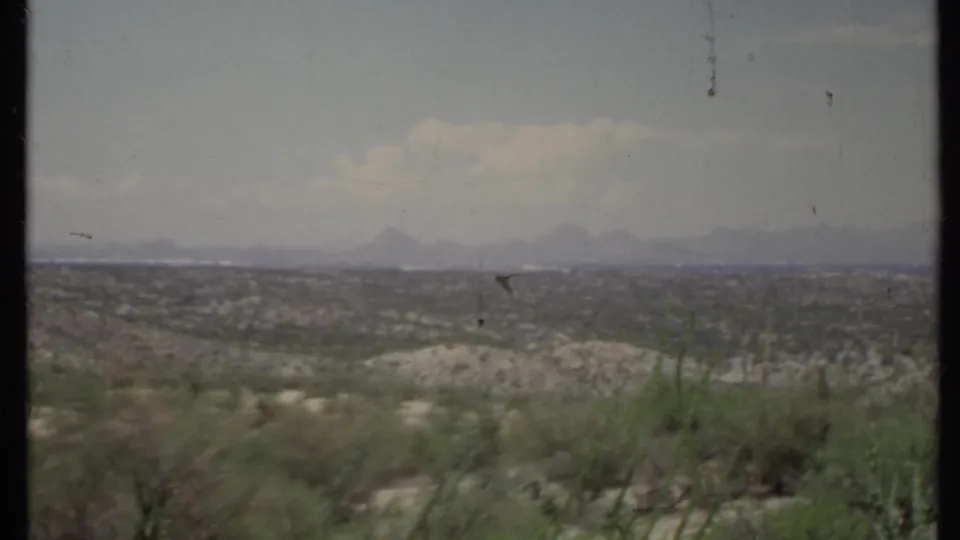 Static 1971 California desert frame showing sparse vegetation across a valley with mountains softened by haze in the distance.