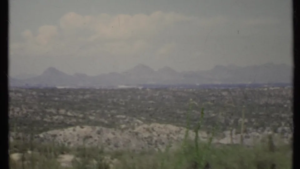 1971 8mm California landscape frame with arid foreground plants, rolling pale hills, and mountain silhouettes across the horizon.