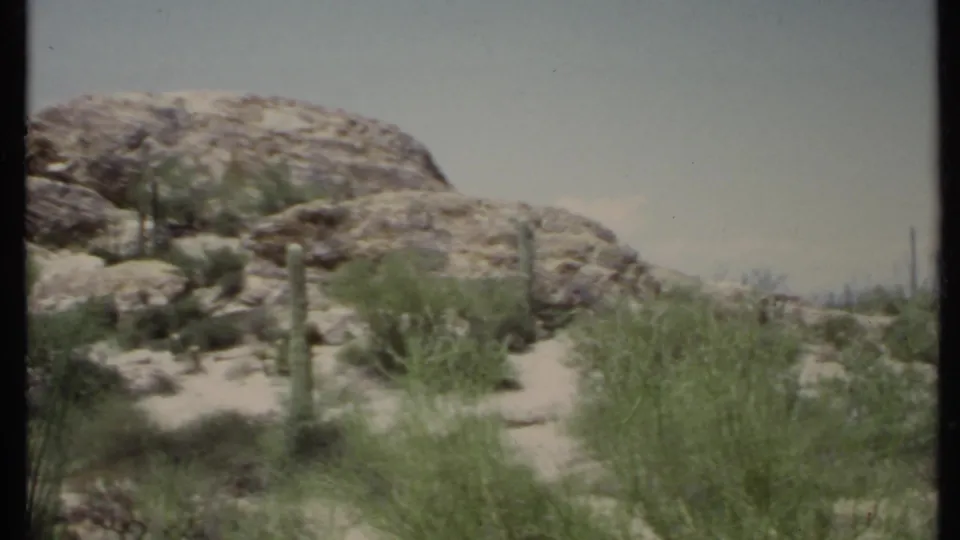 1971 8mm California desert hillside with rocky outcrops, columnar cacti, low shrubs, pale soil, and washed blue sky.