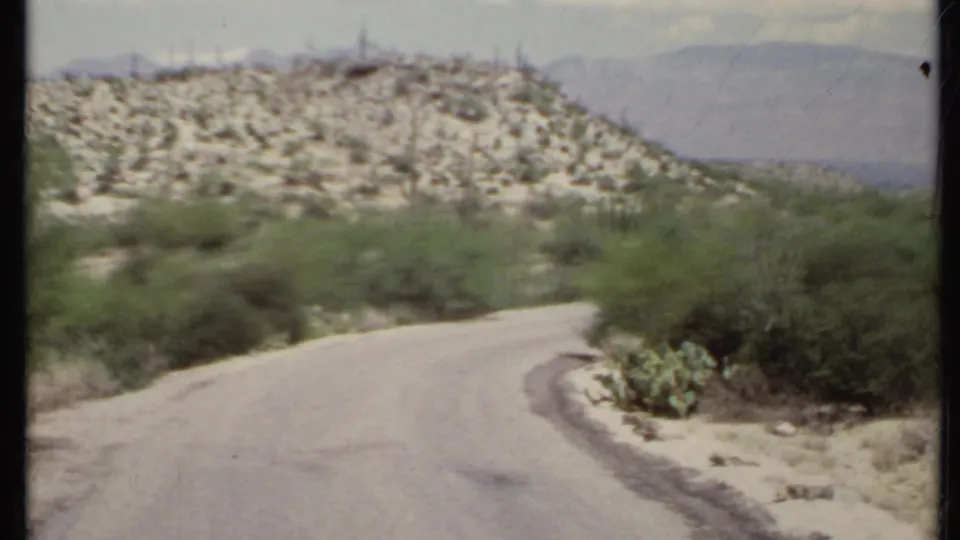 1971 California desert road curving past cacti, shrubs, rocky hills, and hazy mountains in a soft 8mm frame.