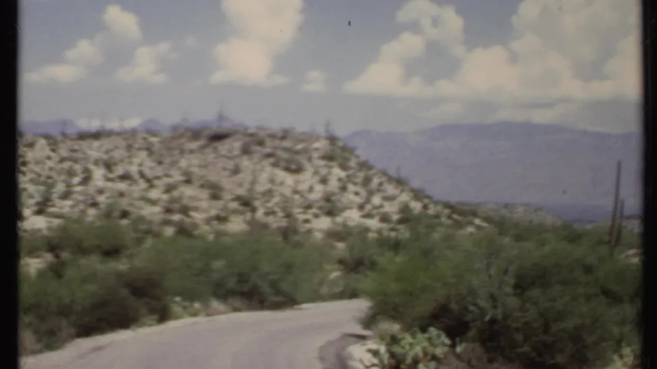 Soft 8mm view of a rocky California desert hill, roadside brush, a curving road, and distant mountain ridges.