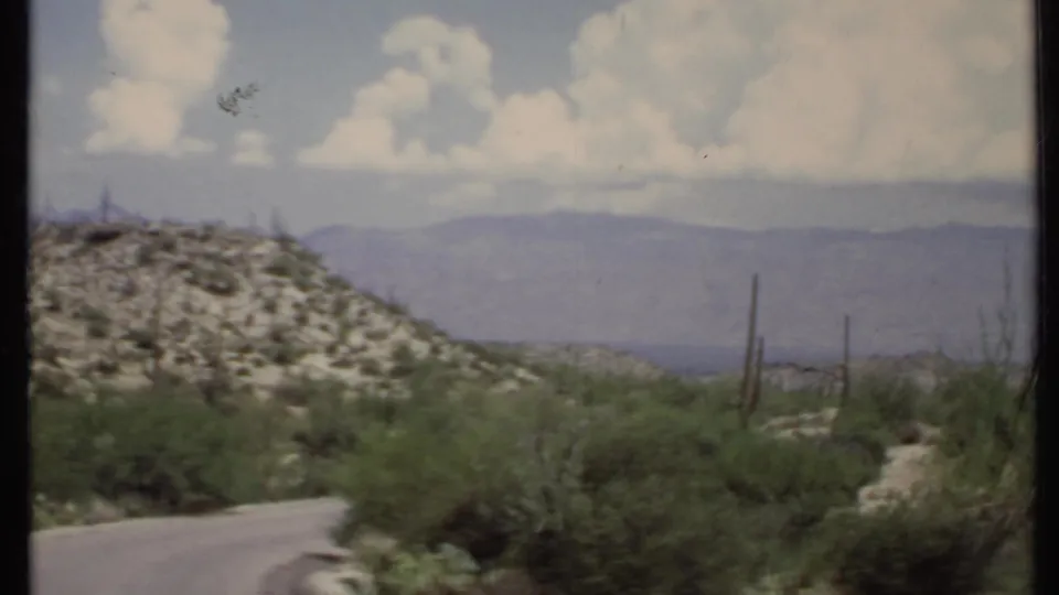 1971 California desert landscape with shrubs, cactus shapes, a road edge, low hills, and mountains under partly cloudy sky.