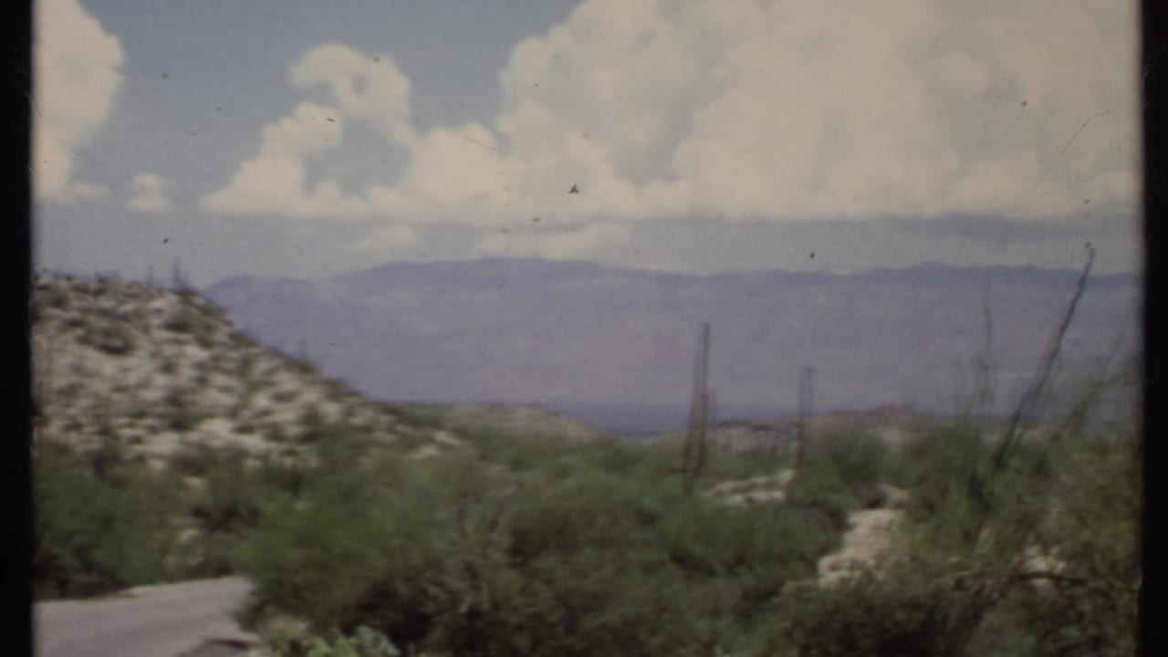 Desert valley road with scrub, rocky hillside, distant mountains, and large clouds.