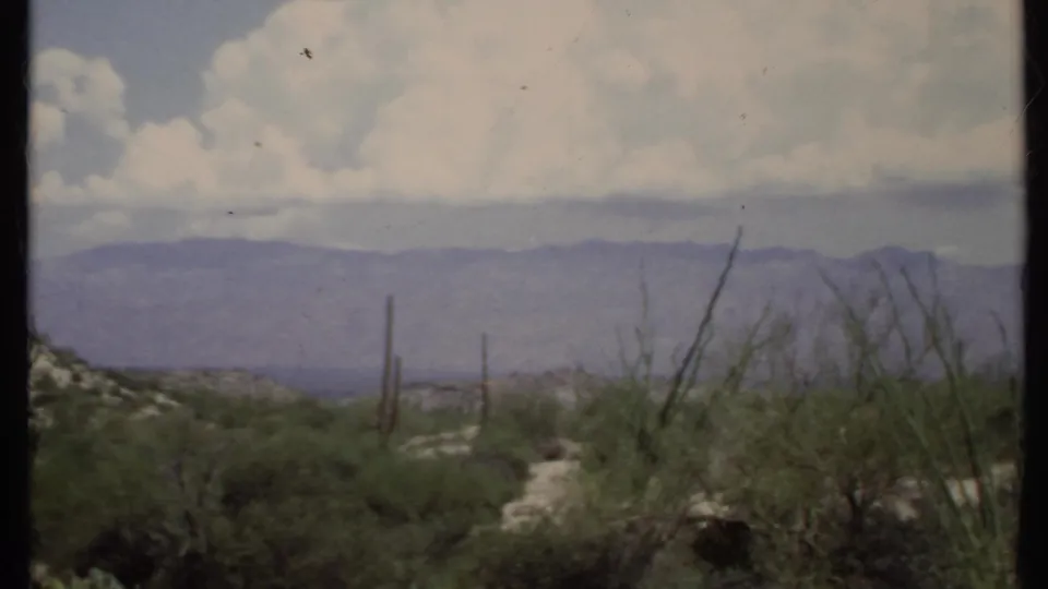 1971 California home-movie frame with cactus-like desert plants, low scrub, layered mountains, and a bright cloud bank across the sky.