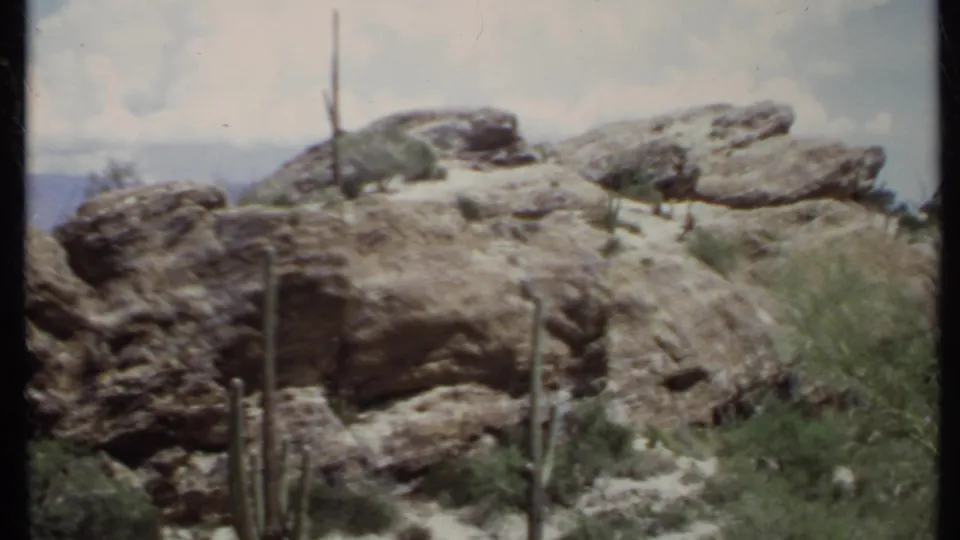 1971 8mm California desert frame showing weathered rock ledges, upright cacti, scrub vegetation, distant hills, and faded color.