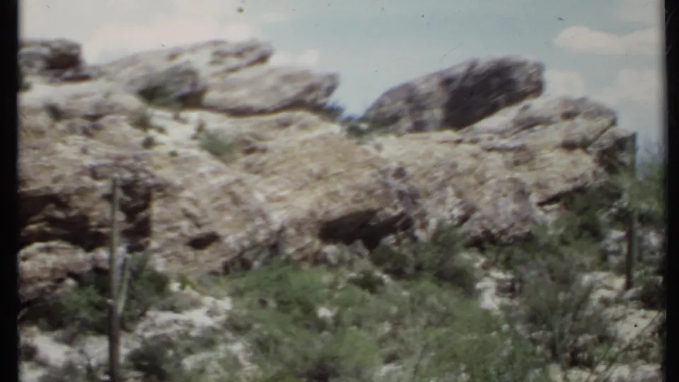 1971 8mm California desert footage frames broad rock outcrops, cactus stems, scrub plants, sandy terrain, and soft sky.