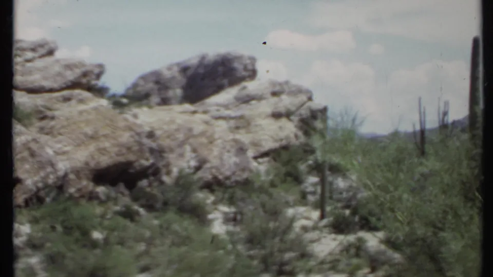1971 8mm California desert view with rocky foreground, upright cacti, green scrub, distant low hills, and faded home-movie color.