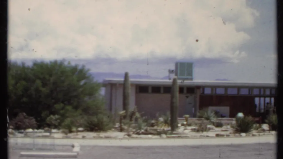 1971 California 8mm frame showing a low modern house, flat roof, front desert planting, tall cacti, and cloudy sky.
