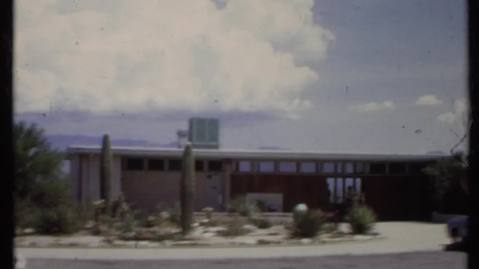 1971 California home-movie frame of a flat-roof house with desert planting, driveway, and partial vehicle at the right edge.