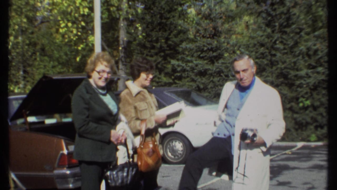 In 1979 Ithaca, three adults stand near parked cars while a man in a white coat holds a camera.