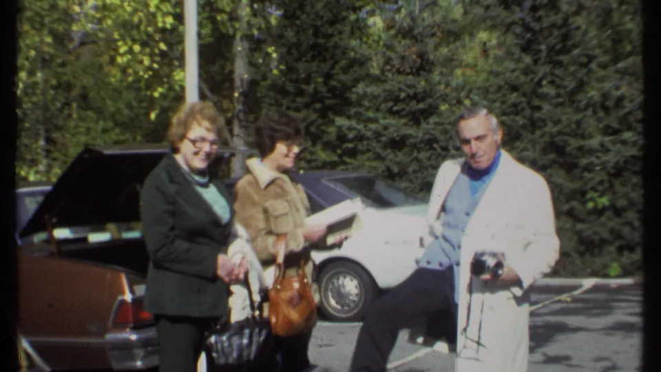 In 1979 Ithaca, three adults stand near parked cars while a man in a white coat holds a camera.