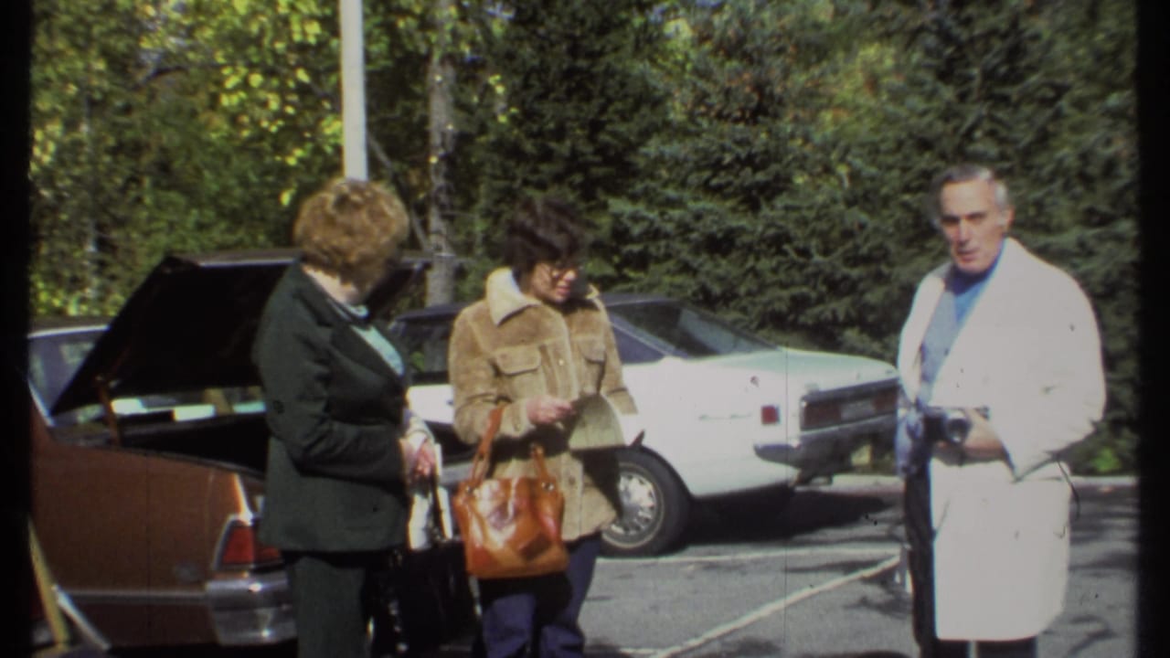 Two women look down near a car while a man in a white coat holds a camera.