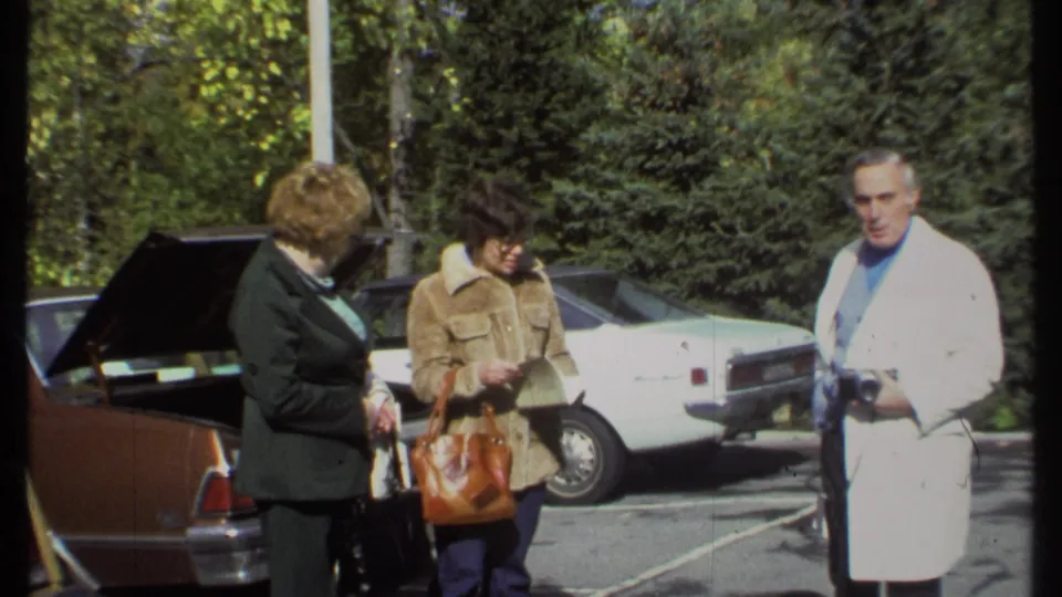 In the Ithaca parking lot, two women stand by a brown car as the man with a camera faces them.