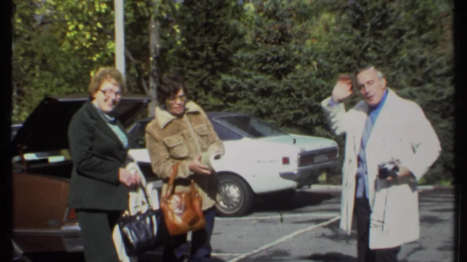 The man in a white coat raises his hand in a dance-like gesture as two women stand in the parking lot.