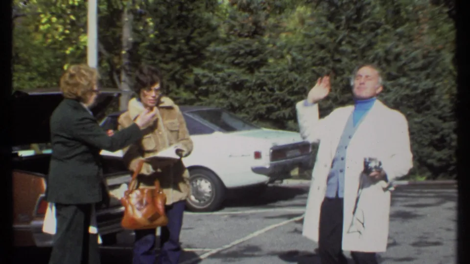 In the final frame, the man with the camera waves or performs while the two women stand near the open car.