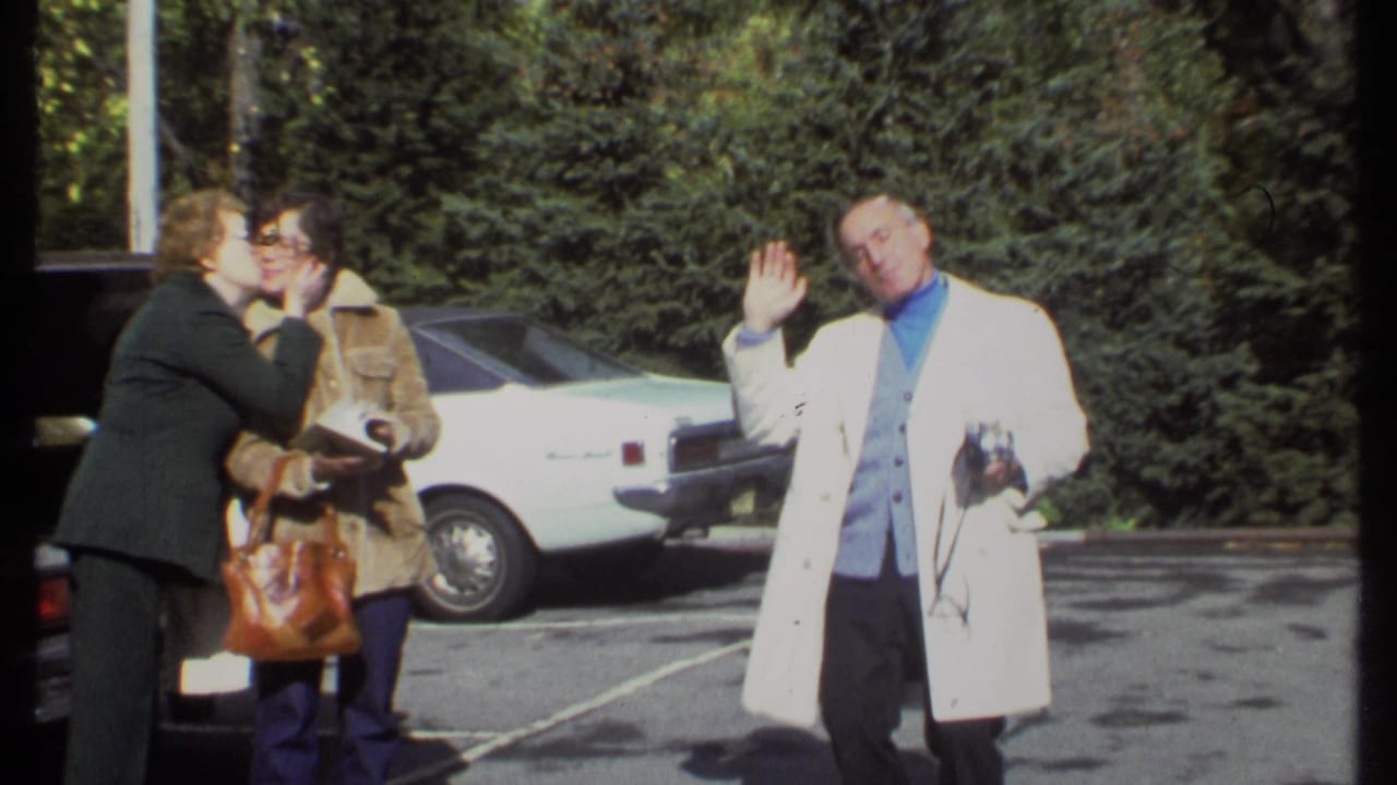 Two women stand close in a parking lot while a man in a white coat waves nearby.