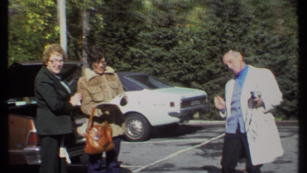 Two women smile near an open car trunk while a man in a white coat stands to the side.