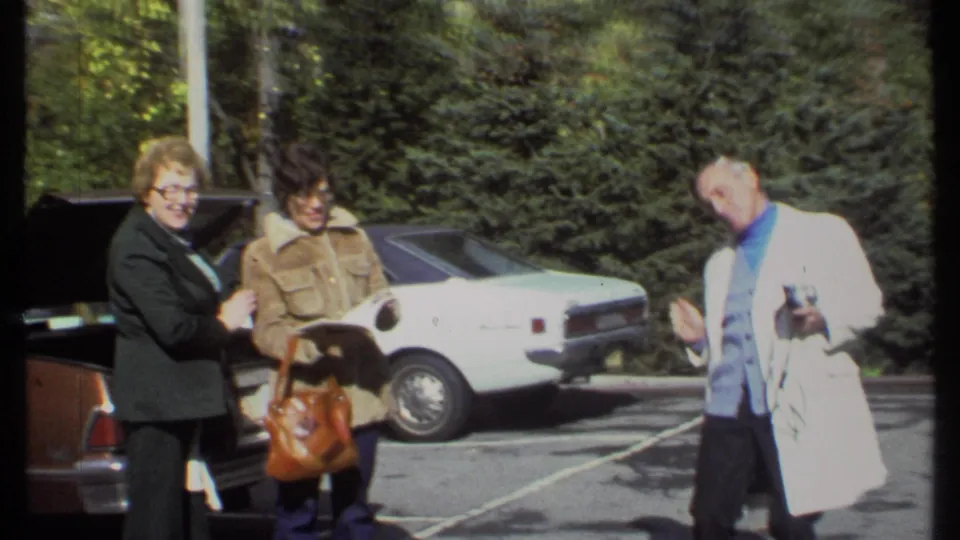 In 1979 Ithaca home-movie footage, two women stand by an open trunk and a white sedan while a man in a white coat gestures at frame right.