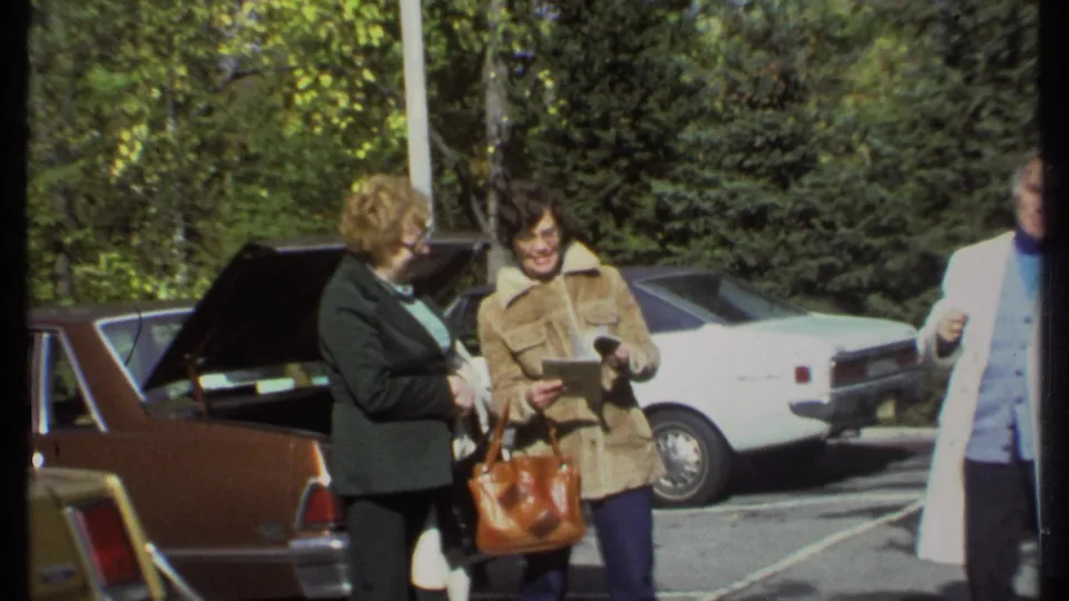 In Ithaca, New York in 1979, two women examine a book or folder near a brown station wagon with its trunk open.