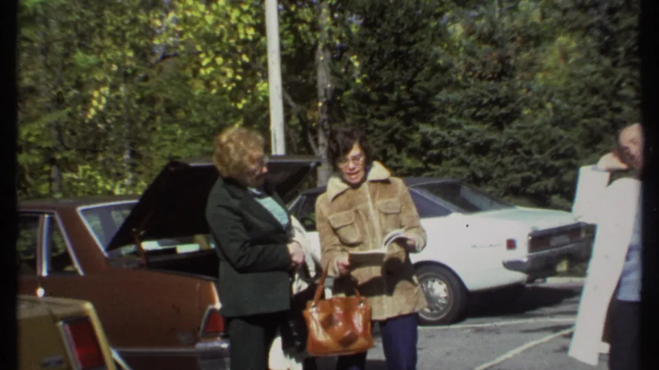 The 1979 Ithaca parking-lot scene shows two women near a brown station wagon and white sedan as the man in a white coat exits the frame edge.