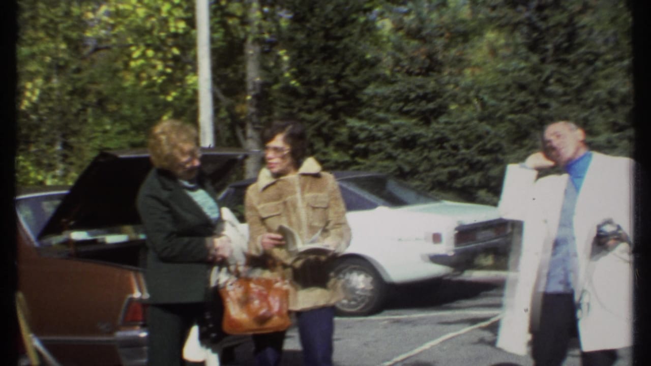 Adults standing near parked cars, with a woman holding a book and a man in a white coat nearby.