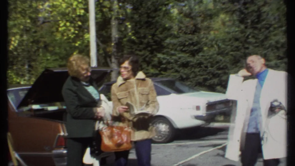 In Ithaca, New York, 1979 home-movie footage, adults stand by parked cars as one woman reads and a man in a white coat gestures.