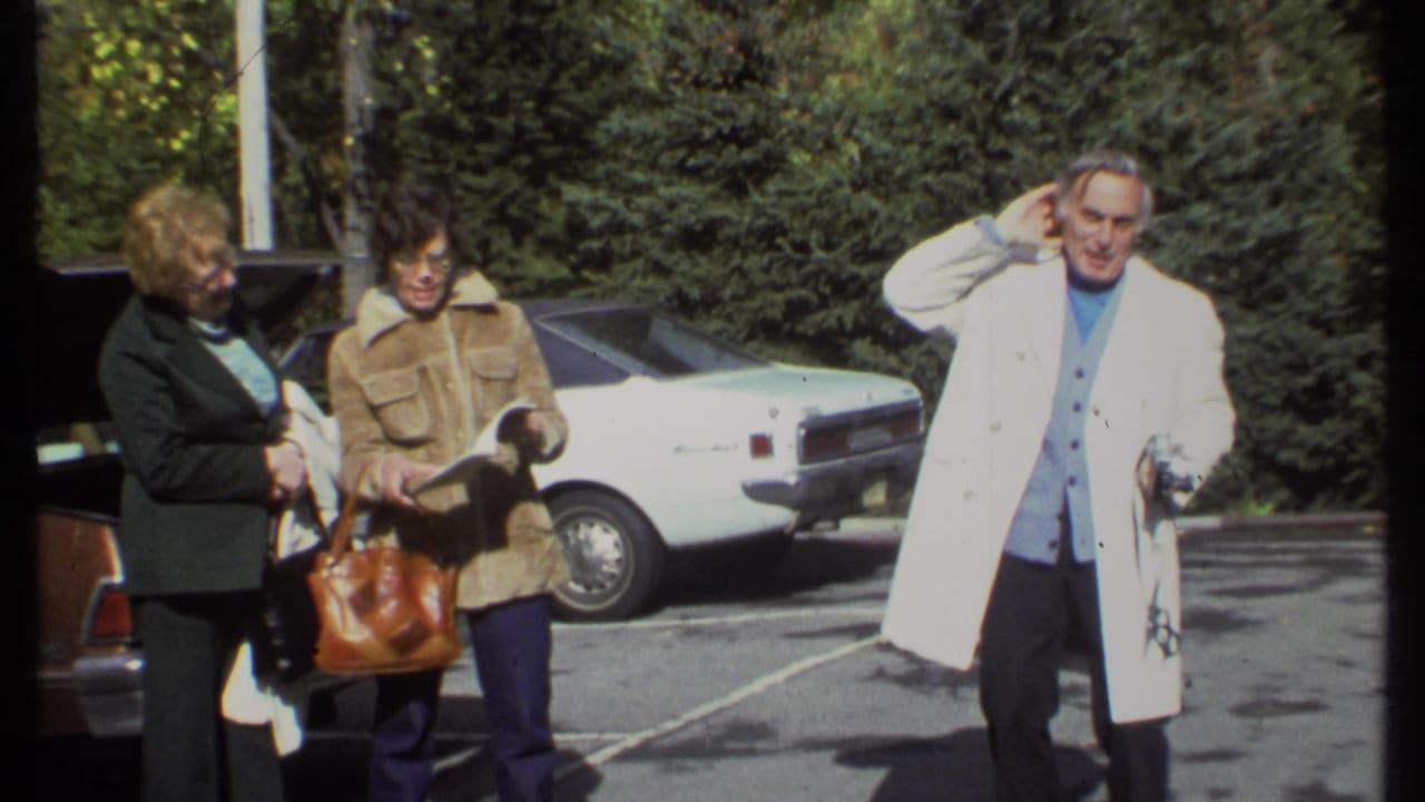 Three adults stand by parked cars while a man in a white coat raises a hand to his head.
