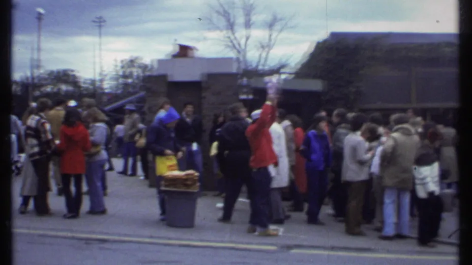 A 1979 Ithaca crowd stands outside a low public building, with adults in coats and one person in red raising an arm.