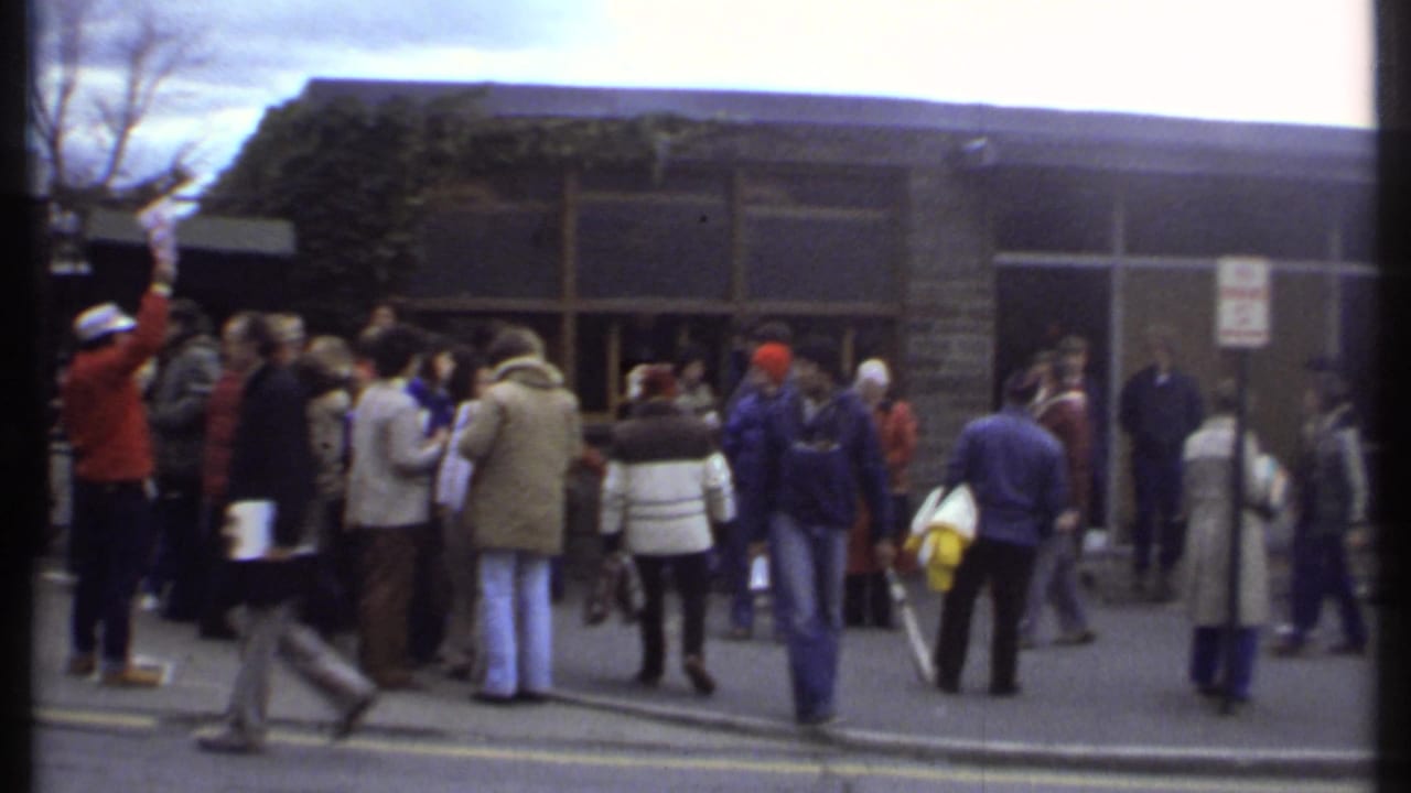 People in coats gather outside a tan brick building near glass doors on an overcast day.
