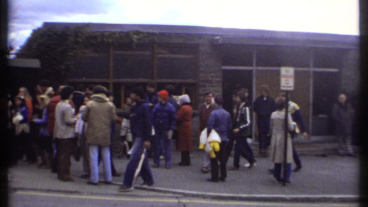 Crowd outside glass doors with a tan building facade and people walking across the foreground.