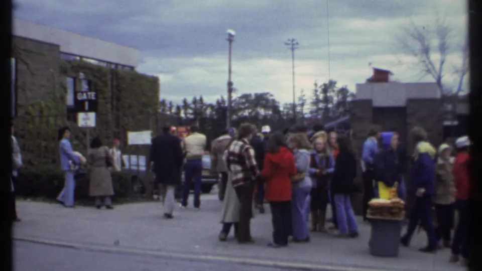 Late-1970s Ithaca home-movie frame showing people gathered near Gate 1 outside a tan building on a cloudy day.