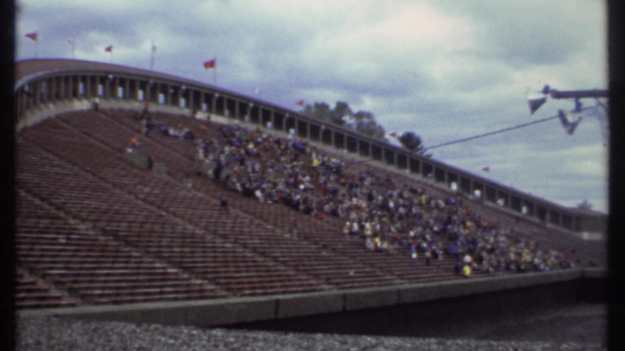 Wide stadium bleachers with scattered spectators under a cloudy sky.