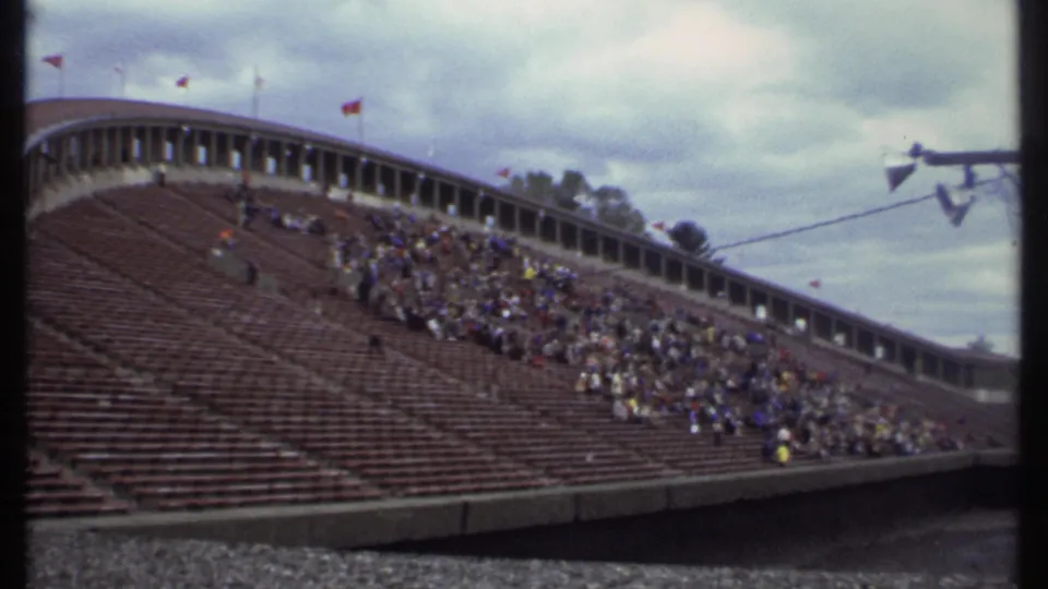 Wide 1979 Ithaca stadium view with red seating, scattered spectators, flags, and a cloudy sky above the stands.