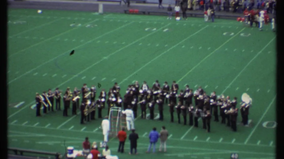 1979 Ithaca football field view with a marching band in formation, sideline observers, yard markers, and green turf.