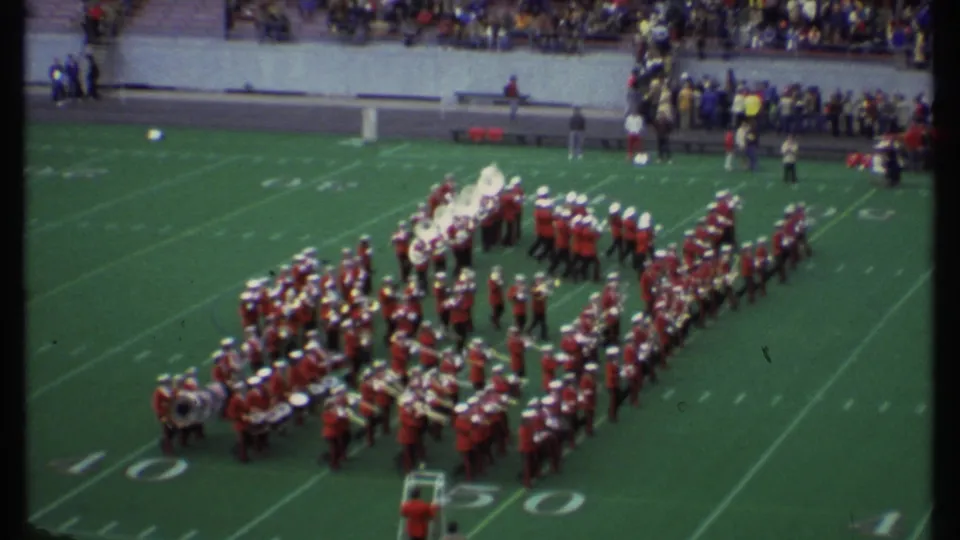 1979 Ithaca, New York 8mm frame showing a red-uniformed marching band in formation on a football field before a game.