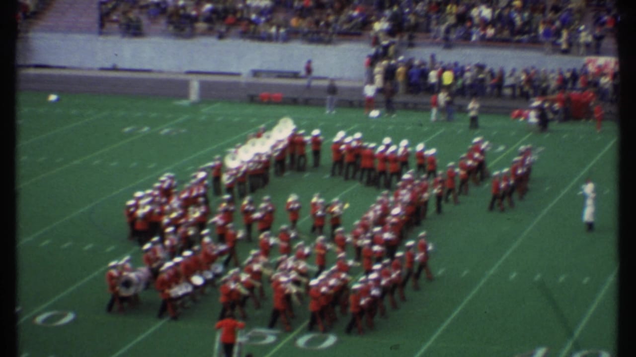 Band members stand in formation near midfield with spectators in the background.